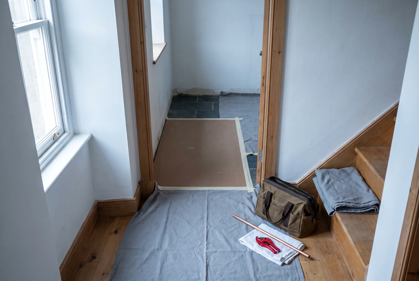 A hallway and bathroom doorway with the floors covered in grey dust sheets and hardboard, a folded sheet on the stair and a closed tool bag by the door.